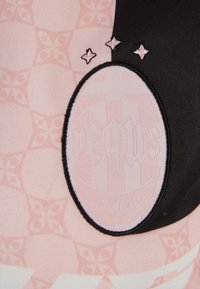 Roza tkanina s geometrijskim vzorcem, ki ima okroglo belo oznako z black besedilom in tremi majhnimi črnimi ter roza zvezdami na strani.