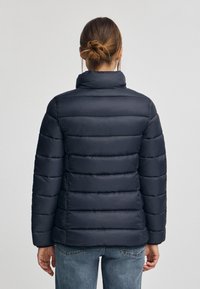 Marinblå puffer-jacka med horisontella quiltade sektioner, hög krage och mjuk textur. Bakvy visar en åtsittande form och elastiska ärmslut.