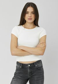 T-shirt blanc à manches courtes côtelé avec un col rond, associé à un jean en denim noir taille haute. Le modèle se tient bras croisés.
