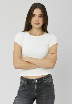 T-shirt blanc à manches courtes côtelé avec un col rond, associé à un jean en denim noir taille haute. Le modèle se tient bras croisés.
