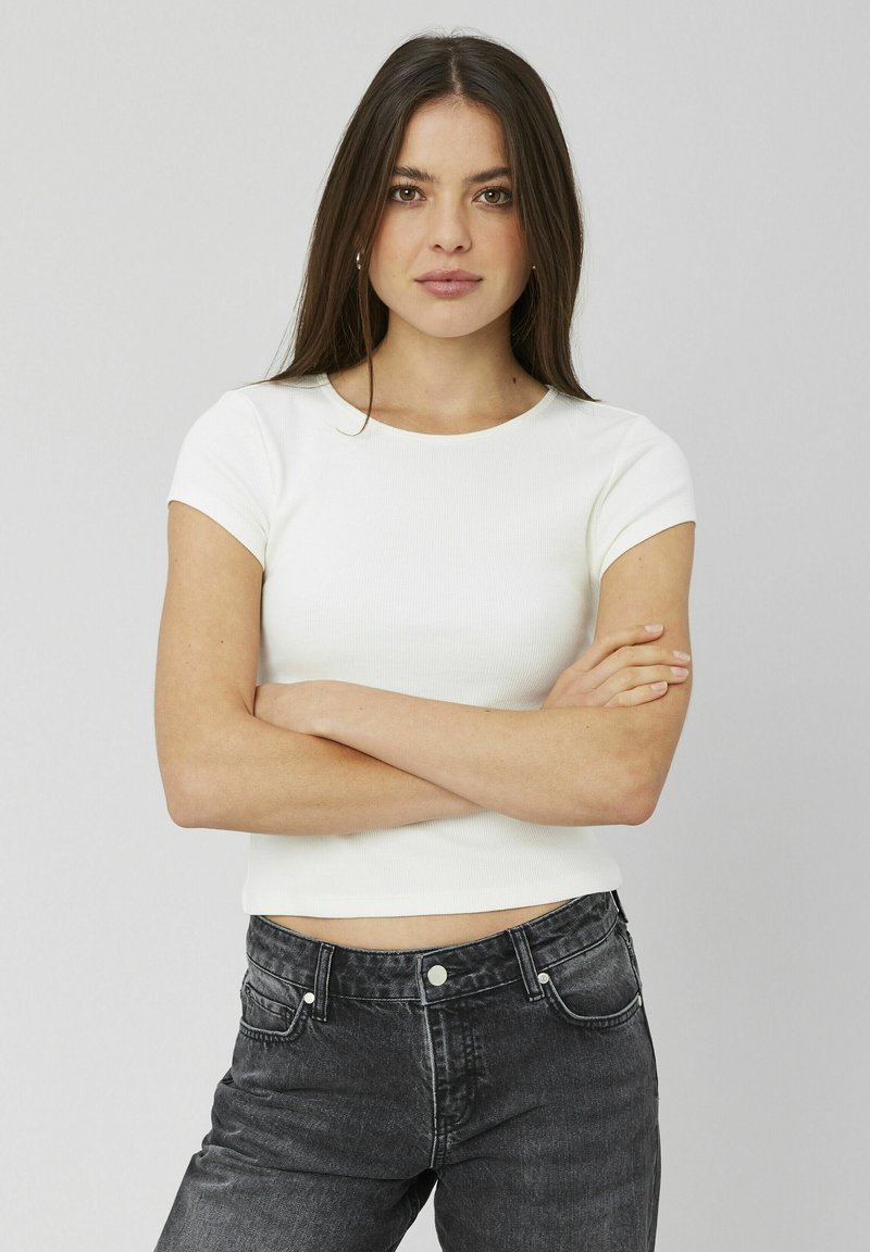T-shirt blanc à manches courtes côtelé avec un col rond, associé à un jean en denim noir taille haute. Le modèle se tient bras croisés.