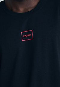 Fekete pamut póló kerek nyakkal. Az elején egy piros téglalap alakú logó található, amelyen a "HUGO" felirat áll.