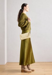 Cardigan en vert olive texturé sur une jupe longue en satin de teinte similaire, associé à des bottes à talons beiges et un sac en paille tressée.