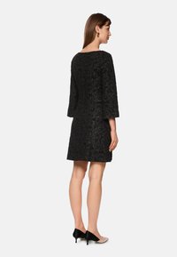 Schwarzes Spitzenkleid mit Dreiviertelärmeln, taillierter Form und floralem Muster, kombiniert mit schwarzen und hautfarbenen zweifarbigen Absätzen.