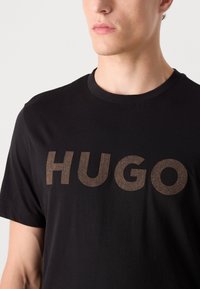 Melns T-krekls ar lielu herringbone raksta "HUGO" logotipu kontrastējošā brūnā krāsā. Apaļš kakla izgriezums un īsas piedurknes.