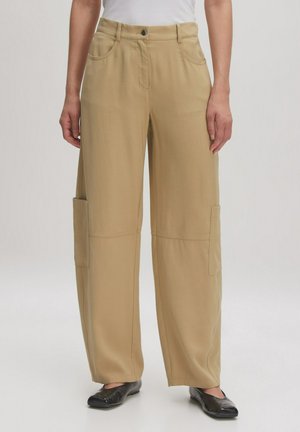 Beige weit geschnittene Hose aus weichem Stoff mit vorderen Taschen und glatter Oberfläche. Kombiniert mit schwarzen flachen Schuhen.