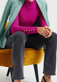 Różowy prążkowany golf z złotymi guzikami, zestawiony z szarymi dżinsami. Modelka siedzi na musztardowo-żółtym fotelu, ubrana w zielonkawy turkusowy żakiet.