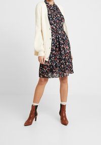 Robe noire à fleurs avec manches courtes et ceinture à la taille, portée sous un cardigan en tricot crème. Bottines à talons bruns avec accents noirs.