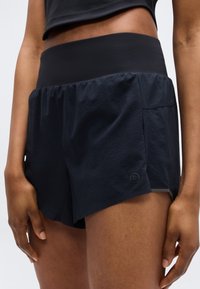 Μαύρο αθλητικό σορτς με φαρδύ, ελαστικό ζωνάρι μέσης, ελαφρύ ύφασμα, υφή στην επιφάνεια και μικρό κυκλικό λογότυπο στο πλάι.