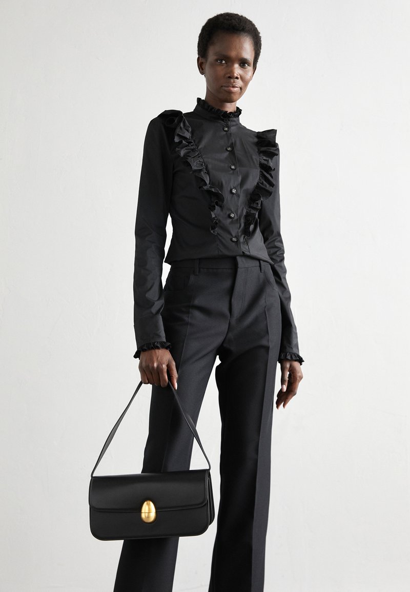 Čierna blúzka s volánmi a gombíkovým zapínaním, spárovaná s prispôsobenými čiernymi nohavicami. Drží štruktúrovanú čiernu kabelku s zlatým akcentom.
