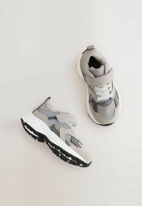 Szare i białe dziecięce buty sportowe z siateczkowymi i zamszowymi detalami, sznurówkami oraz rzepem na jednolitym jasnym tle.