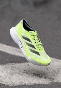 Chaussure de course jaune vif avec des rayures noires, flottant au-dessus d'un bitume avec des marquages routiers blancs et de petits grains de gravier.