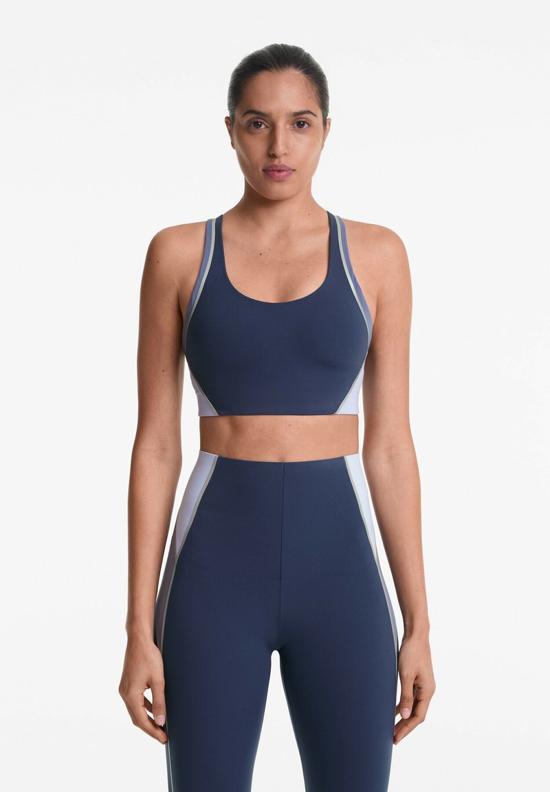 Sportbeha in marineblauw met contrasterende lichtblauwe en grijze zijkanten. Glad, rekbaar materiaal met een ronde halslijn en brede banden.