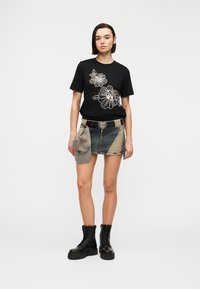 Femme aux cheveux courts et foncés portant un t-shirt noir à fleurs, une mini-jupe en jean bicolore avec ceinture en forme de cœur, des bottes de combat noires, tenant une casquette grise à côté.