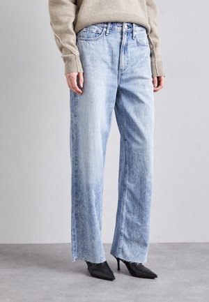Personne portant un jean large bleu clair aux ourlets effilochés, un pull beige et des chaussures à talons hauts pointues noires sur un fond gris uni.