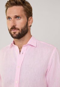 Camisa de lino color rosa claro con una textura suave, cuello de botón y una sola tapeta de botones en el pecho. Presenta un patrón de tejido sutil.