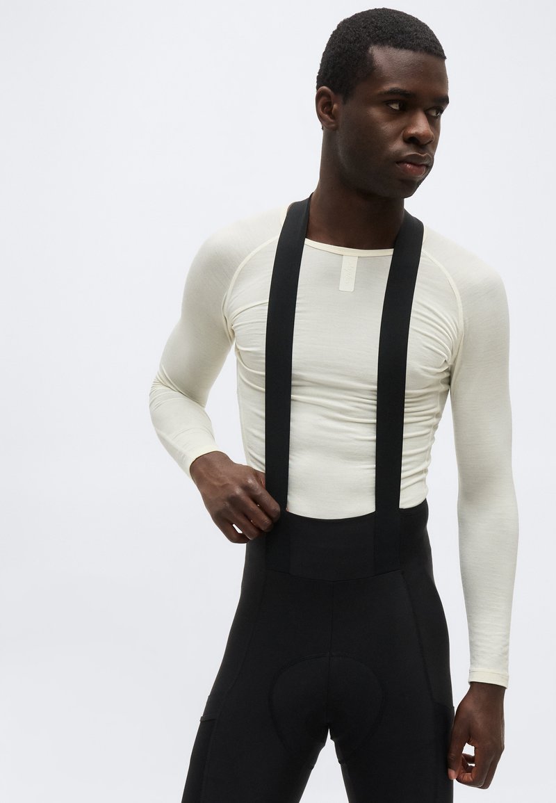 Man wearing a fitted long-sleeve cream shirt and black cycling bib pants with suspenders against a plain white background.