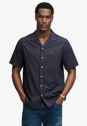 Mann trägt ein dunkles kurzärmeliges Hemd mit Knöpfen und blaue Jeans, steht mit Händen in den Taschen vor einem schlichten Hintergrund.