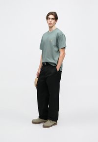 Hellgrünes Baumwoll-T-Shirt mit kleinem gelben Logo, kombiniert mit schwarzen weiten Hosen und grünen Schuhen mit strukturierter Oberfläche.