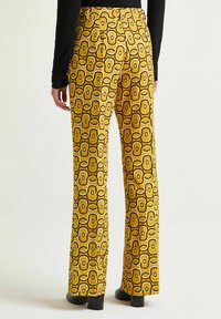Pantaloni svasati realizzati in tessuto giallo lucido, caratterizzati da un motivo geometrico rétro di forme incrociate in marrone e crema.