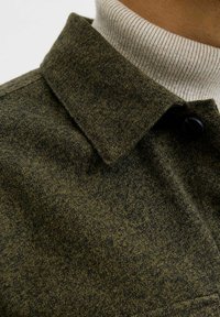 Nærbillede af en person iført en mørkegrøn struktureret frakke over en beige ribbet turtleneck sweater.