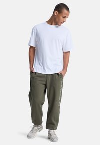 Vit bomullströja i kombination med olivgröna sweatpants med logotryck, fulländat med ljusgrå sneakers, stående i en avslappnad pose.