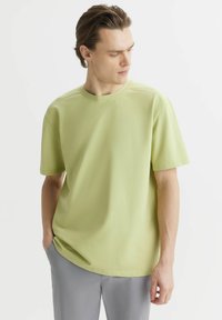 Jasnozielona koszulka z krótkim rękawem, wykonana z miękkiej, teksturowanej tkaniny, z okrągłym dekoltem i luźnym krojem, zestawiona z szarymi spodniami.