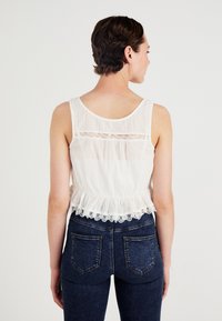 Witte mouwloze top met kantafwerking, gerimpelde onderrand en doorschijnende panelen aan de achterkant. Gecombineerd met donkerblauwe spijkerbroek.