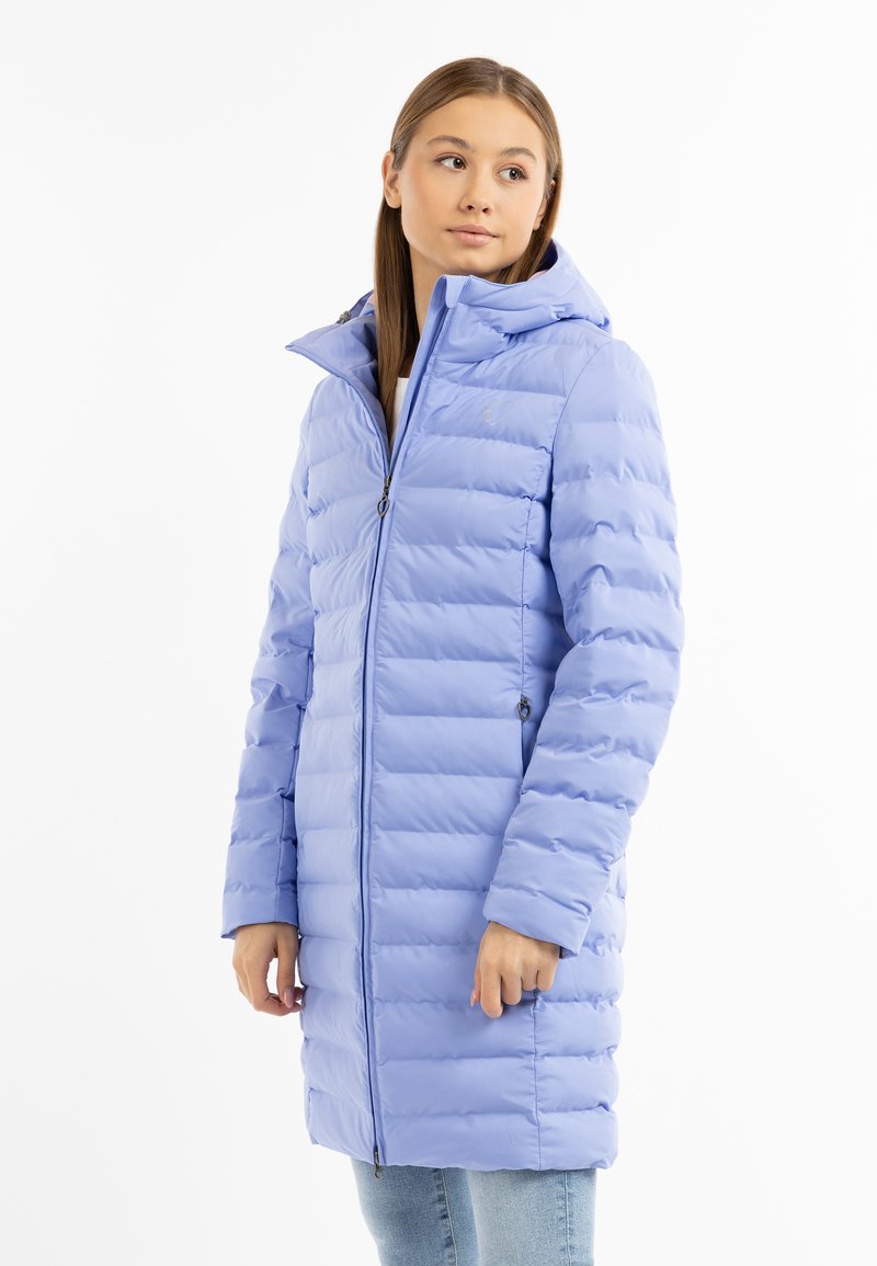 Giovane donna che indossa un cappotto trapuntato con cappuccio di un leggero colore viola, con chiusura frontale a zip, in piedi davanti a uno sfondo bianco.