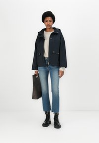Veste navy avec capuche, quincaillerie dorée, deux poches avant, portée sur un pull beige, associée à un jean flare bleu clair et des bottes noires.