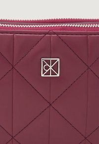 Pungă matlasată burgundy cu cusături diagonale, detalii cu logo argintiu și închidere cu fermoar. Textură netedă, design compact.