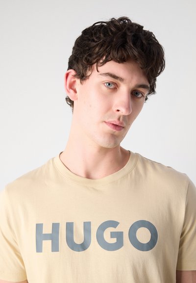 Бежова памучна тениска с голям сив логотип "HUGO" отпред. Тениската има кръгло деколте и къси ръкави.