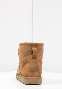 Bottine en suedine beige avec une texture douce, une forme arrondie et une petite languette à l'arrière. Présente un patch en cuir avec une marque sur le talon.