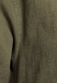 Tela de color verde oliva con un tejido texturizado, mostrando una costura cosida y sutiles acentos negros. La tela parece suave y ligeramente áspera.