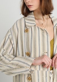 Pruhovaná bunda v krémové barvě s modrými a žlutými pruhy, s límcem, velkými knoflíky a širokými rukávy; texturovaná tkanina.