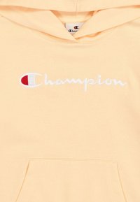 Sudadera con capucha color melocotón y un bolsillo frontal. Presenta el texto bordado "Champion" en blanco con un acento rojo y azul. Textura suave.