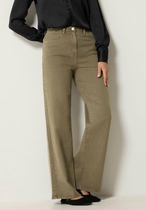 Person trägt hoch taillierte, weit geschnittene olivgrüne Hosen, eine schwarze Langarmbluse und schwarze Ballerinas und steht vor einem neutralen Hintergrund.