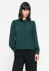 Teal knappskjorte med krage, lange ermer og elastiske mansjetter; har et teksturert stoff og knapper foran for lukking.