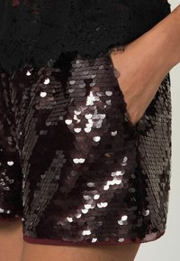 Burgundi színű flitteres rövidnadrág fényes, texturált felülettel rendelkezik, oldalsó zsebbel és egyszínű szegéllyel az alján. A felső része fekete csipke.
