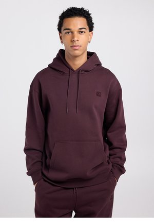 Jonge man met kort krullend haar, gekleed in een donkerbordeauxrode hoodie en bijpassende joggingbroek, staand met handen in de zakken tegen een effen achtergrond.