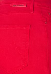Jeans de mezclilla rojo brillante que muestran el bolsillo trasero con un detalle de costura en forma de "M" y una etiqueta de cuero de marca que dice "MOTHER" encima del bolsillo.
