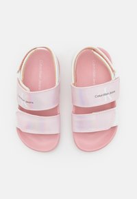 Vaaleanpunaiset sandaalit, joissa on kaksi säädettävää hihnaa, valmistettu hohtavasta materiaalista. Tekstuuripohjassa on Calvin Klein Jeans -brändin logo hihnoissa.