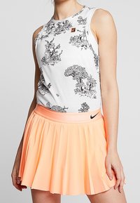 Femme portant un haut blanc sans manches avec des illustrations noires de la nature et une jupe de tennis plissée couleur pêche de Nike.