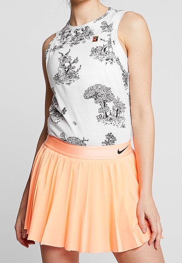 Femme portant un haut blanc sans manches avec des illustrations noires de la nature et une jupe de tennis plissée couleur pêche de Nike.