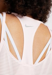 Canottiera sportiva rosa chiaro con una texture a rete e spalline blu, con un piccolo logo Nike grigio sulla parte superiore della schiena.