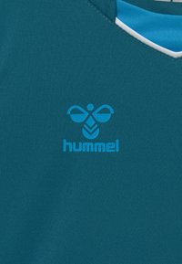 T-shirt de sport teal avec un design en v. Le tissu texturé affiche un logo Hummel bleu au centre avec un élément graphique au-dessus.