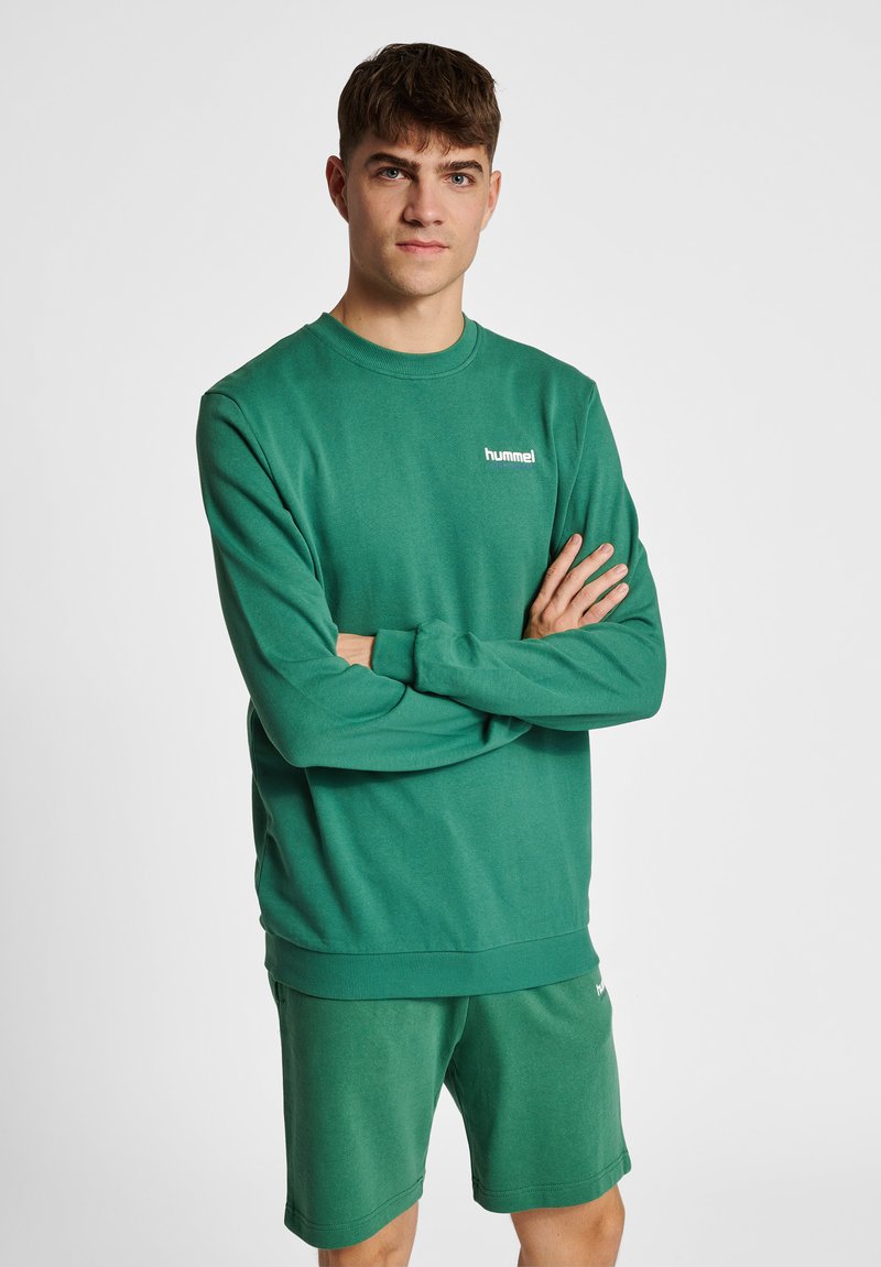 Ung mand med kort brunt hår står med armene krydset iført en grøn hummel sweatshirt og matchende grønne shorts.