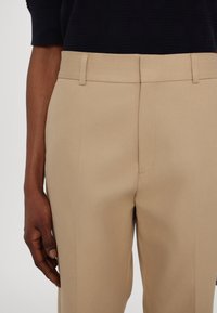 Section médiane d'une personne portant un pantalon beige sur mesure et un haut noir, la main détendue sur le côté, sur un fond uni.