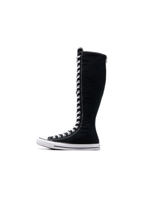 Bottine sneaker noire montante avec lacets blancs s’étendant de la pointe jusqu’à juste en dessous du genou, semelle blanche et coutures contrastantes.