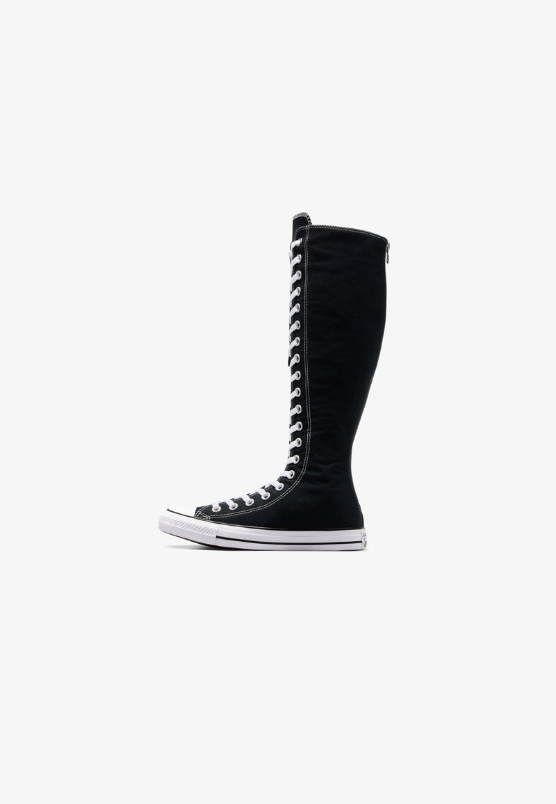 Bottine sneaker noire montante avec lacets blancs s’étendant de la pointe jusqu’à juste en dessous du genou, semelle blanche et coutures contrastantes.